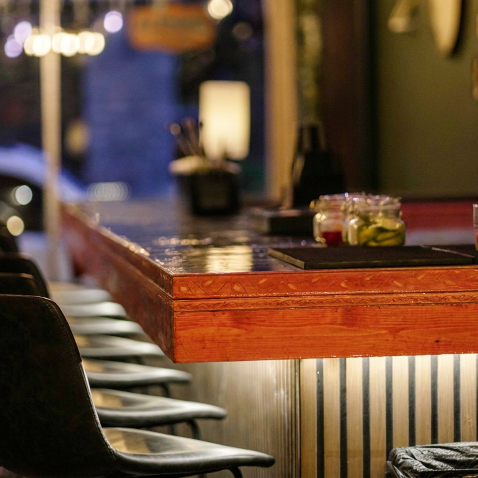 Warmly lit restaurant bar with wooden countertop, black stools, small condiment jars, and a drink against a windowed backdrop.